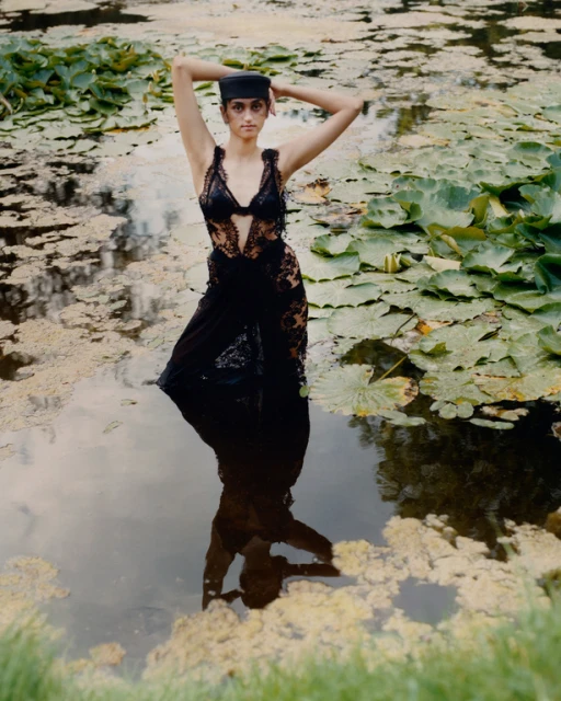 Woman in a black dress and hat stands in a pond with lily pads, reflected in the water, outdoor portrait, natural light, editorial fashion.