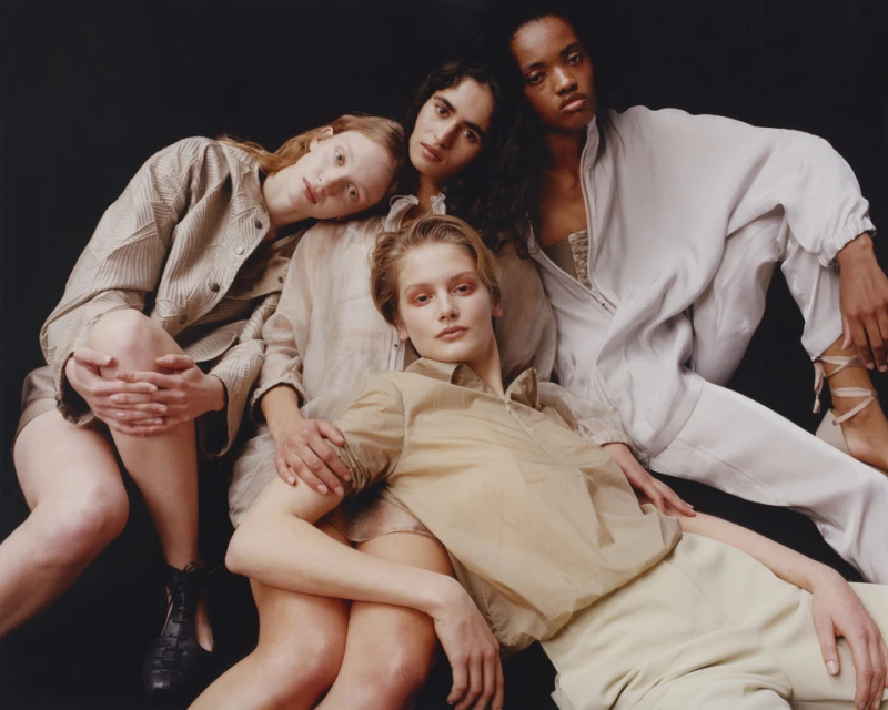 Four diverse female models in a tonal fashion shoot. They wear neutral-colored outfits in a studio setting with dark backdrop.