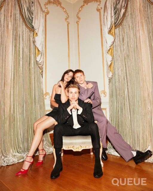Three models pose indoors in a studio with cream-colored walls and gold accents. A woman and man stand behind a seated man.