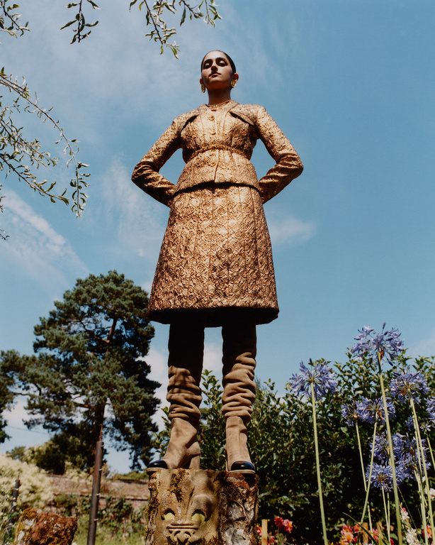 A fashionable woman in a gold suit stands tall on a stone pedestal outdoors, with a bright blue sky and trees in the background.