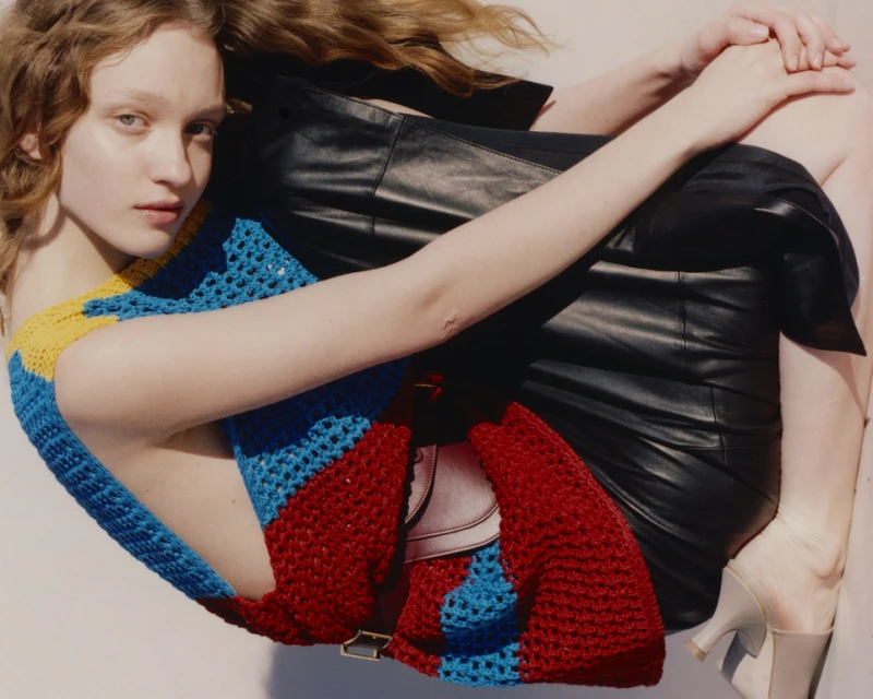 Indoor shot of a young adult woman with long wavy hair wearing a colorful knit top and a leather skirt posing against a white background.