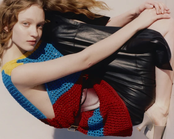 Indoor shot of a young adult woman with long wavy hair wearing a colorful knit top and a leather skirt posing against a white background.