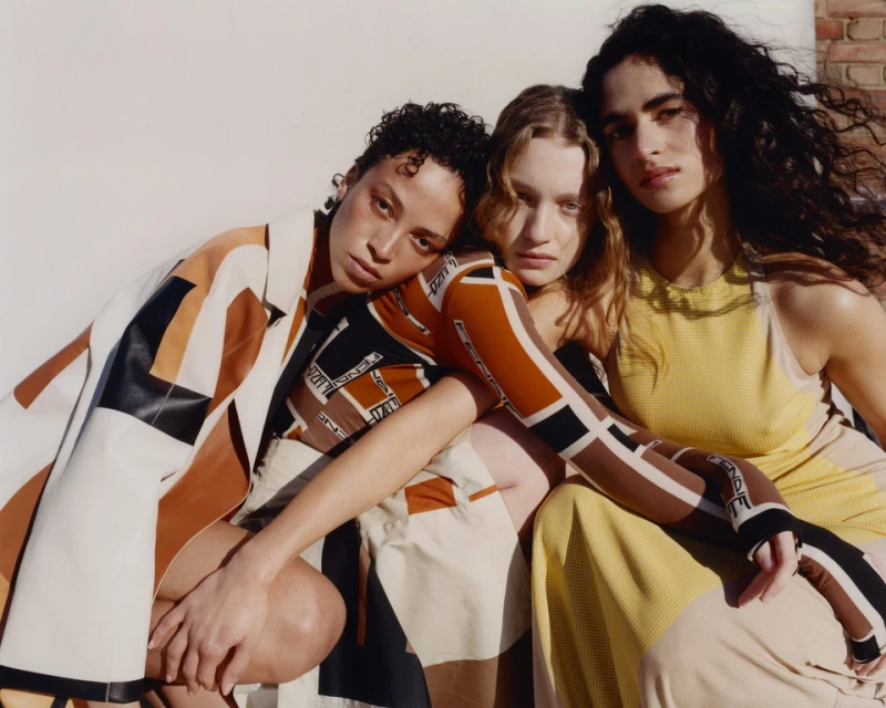 Three diverse young women in fashion apparel pose in a close studio shot. Geometric patterned tops and dresses in earth-tone colors.
