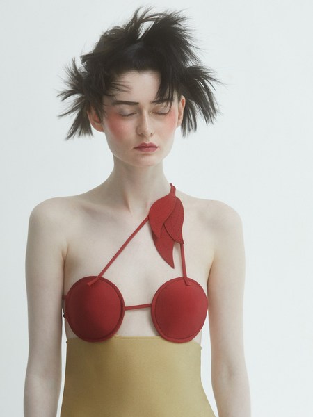 A fair-skinned woman with dark hair and closed eyes poses in a cherry-themed garment against a bright white background.
