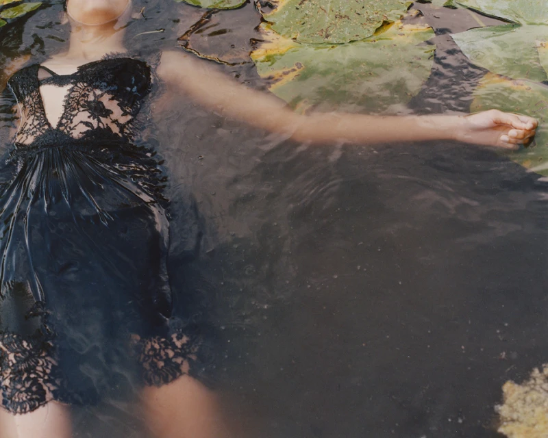Woman in black lace dress floating in a pond with lily pads, captured in a dreamy, ethereal style with soft, diffused lighting.