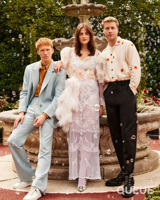 Three fashionably dressed people pose in front of a stone fountain in a garden setting with abundant green foliage and floating bubbles.