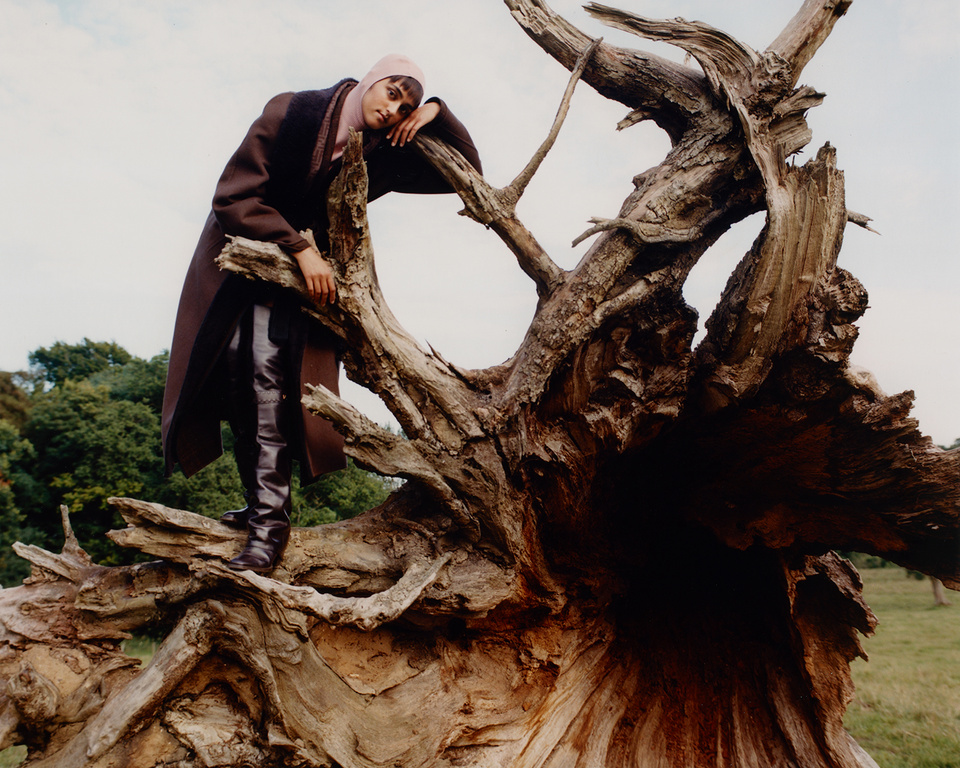 A woman poses on a fallen tree trunk wearing a brown coat, black boots, and pink headscarf against a backdrop of sky and foliage.