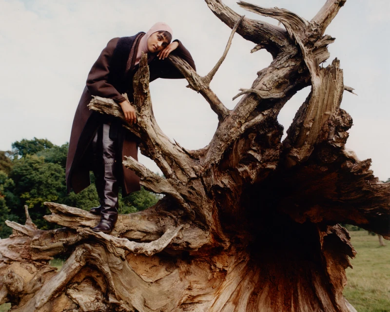 A woman poses on a fallen tree trunk wearing a brown coat, black boots, and pink headscarf against a backdrop of sky and foliage.