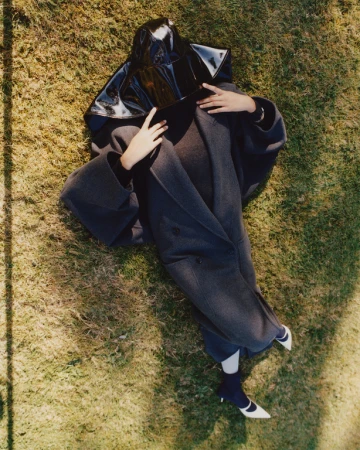 A woman in a gray coat and black vinyl hood rests on green grass, facing the camera. Her feet are in the lower right, and hands are visible.