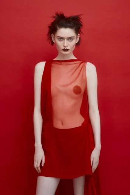 A slender young woman with dark hair and striking makeup stands in front of a seamless red backdrop, wearing a semi-sheer red dress.