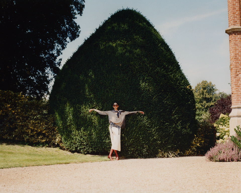 Adult female posing outdoors in a beige outfit in front of a large, rounded bush, with a brick building in the background.