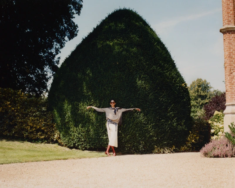 Adult female posing outdoors in a beige outfit in front of a large, rounded bush, with a brick building in the background.
