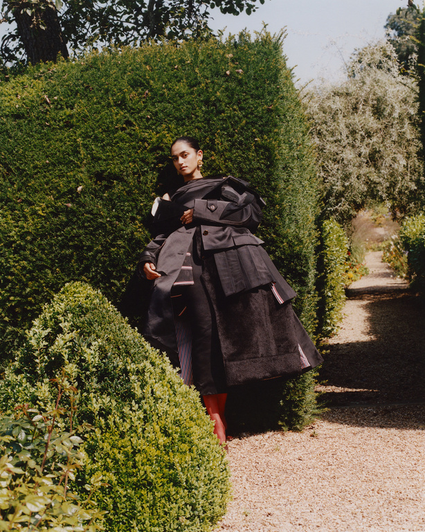 A person in an oversized black outfit poses in a garden setting with trimmed hedges and a gravel path, bathed in soft daylight.
