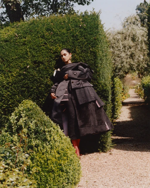 A person in an oversized black outfit poses in a garden setting with trimmed hedges and a gravel path, bathed in soft daylight.