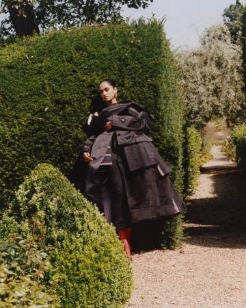 A person in an oversized black outfit poses in a garden setting with trimmed hedges and a gravel path, bathed in soft daylight.