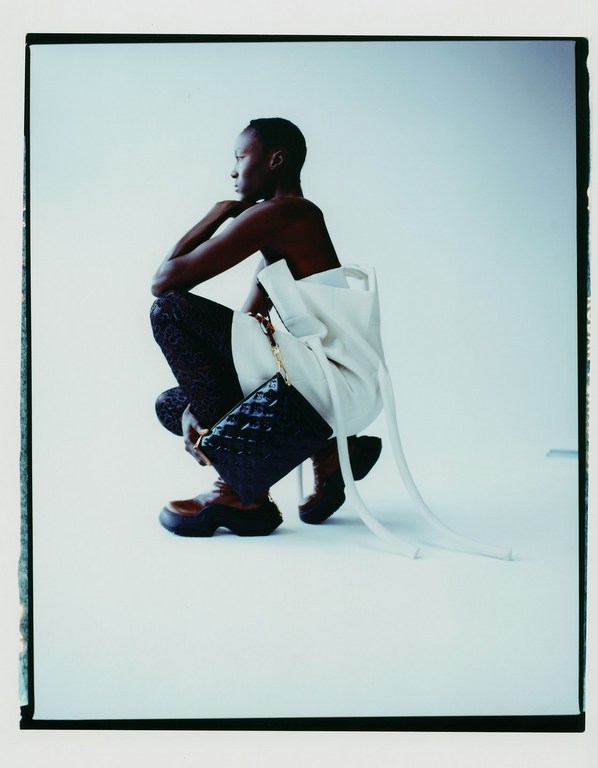 Woman poses in fashion shot against a white background, wearing textured pants, platform boots, a white garment, and carrying a handbag.