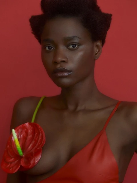 Studio portrait of a black woman with short hair, wearing a red dress, posing in front of a red background with a red Anthurium flower.