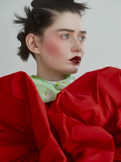 Portrait of a young woman with dark hair and dramatic red lipstick, wearing a red garment against a neutral backdrop.