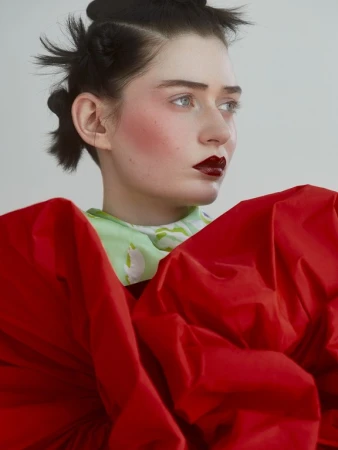 Portrait of a young woman with dark hair and dramatic red lipstick, wearing a red garment against a neutral backdrop.