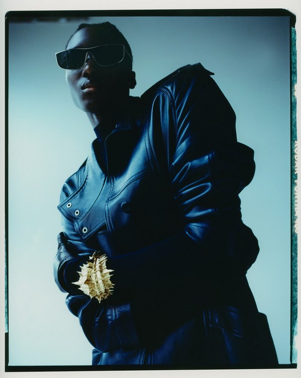 Fashionable person in a leather jacket and sunglasses holds a decorative object against a light blue background in a studio shot.