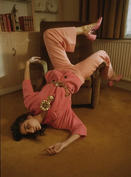 Woman in pink suit poses upside down on a chair in an indoor setting; fashion, style, and creative photography.