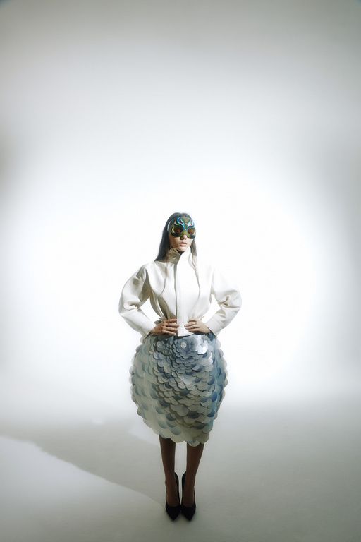 Fashion model poses in a studio wearing a white jacket, sculptural skirt, and futuristic glasses against a neutral backdrop.