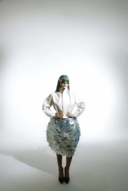 Fashion model poses in a studio wearing a white jacket, sculptural skirt, and futuristic glasses against a neutral backdrop.