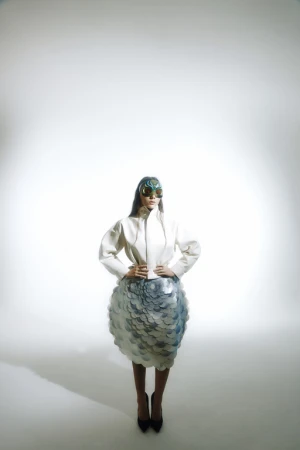 Fashion model poses in a studio wearing a white jacket, sculptural skirt, and futuristic glasses against a neutral backdrop.