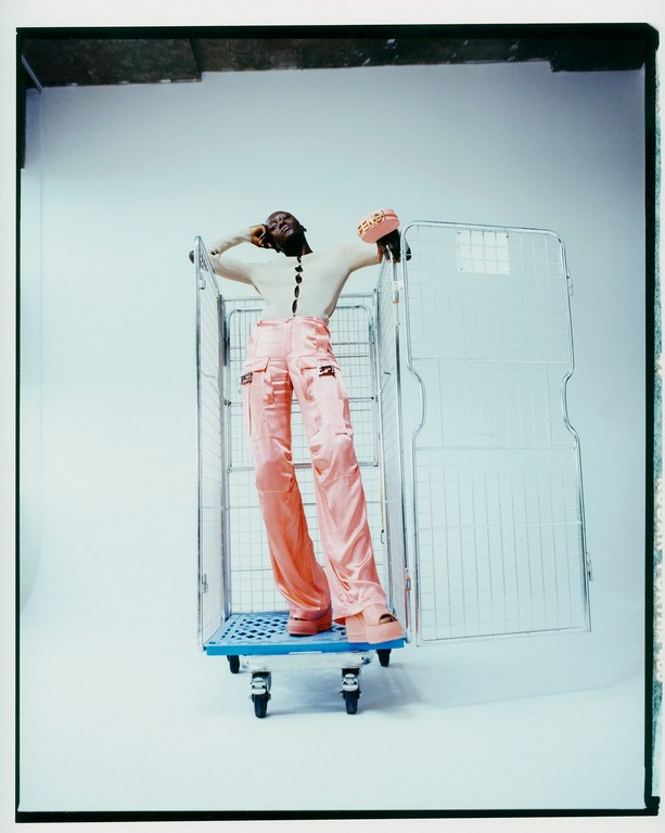 A model poses in a wire shopping cart in a studio. They are wearing a cream top, pink pants, and pink accessories, styled in modern fashion.