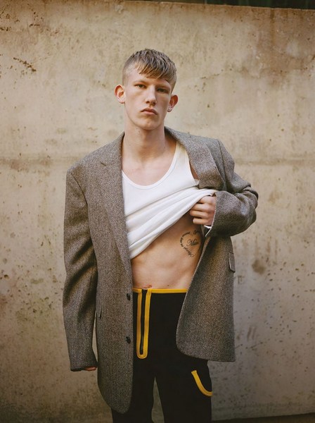 Fashion shot of a young adult man wearing a jacket, white tank top, and pants, showcasing a heart tattoo on his abdomen against a wall backdrop.