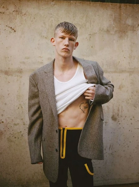 Fashion shot of a young adult man wearing a jacket, white tank top, and pants, showcasing a heart tattoo on his abdomen against a wall backdrop.