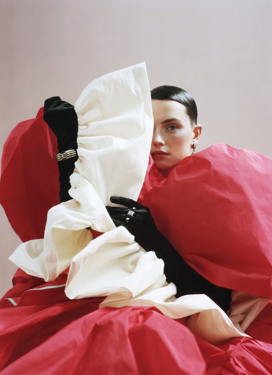 Woman wearing a sculptural red and white gown and long black gloves, striking a pose against a light pink background, portrait.