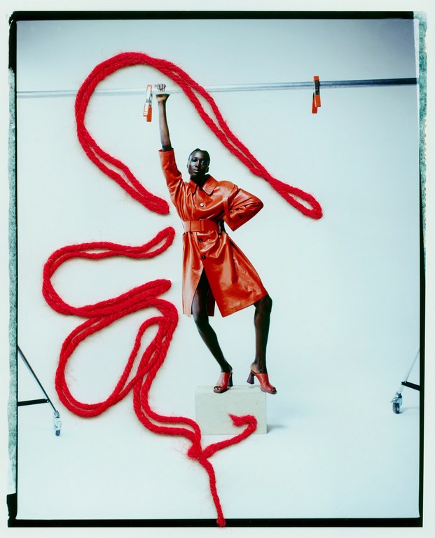 An adult woman poses in a shiny coral coat and red shoes on a white box in a photo studio, with a red yarn prop.