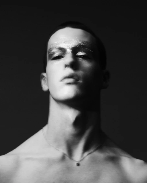 Monochrome portrait of a shirtless person with glitter makeup, against a dark background, showcasing dramatic beauty lighting and style.