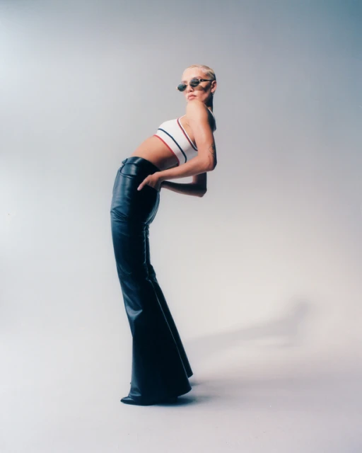 Fashionable adult woman with short blonde hair posing in a crop top and black flared pants in a studio setting with soft lighting.