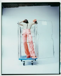 Model standing inside a metal wire roll container wearing pink satin cargo trousers and a cream top styled by Katie Felstead.