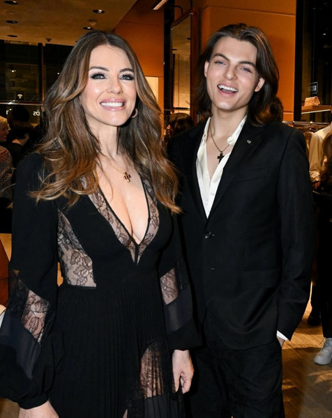 Elegant portrait of a smiling woman in a black lace dress and a young man in a suit, posing at an indoor fashion event.