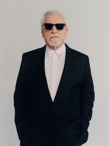 Portrait of older adult male wearing sunglasses and a black suit, posing against a white studio backdrop with hands in pockets.