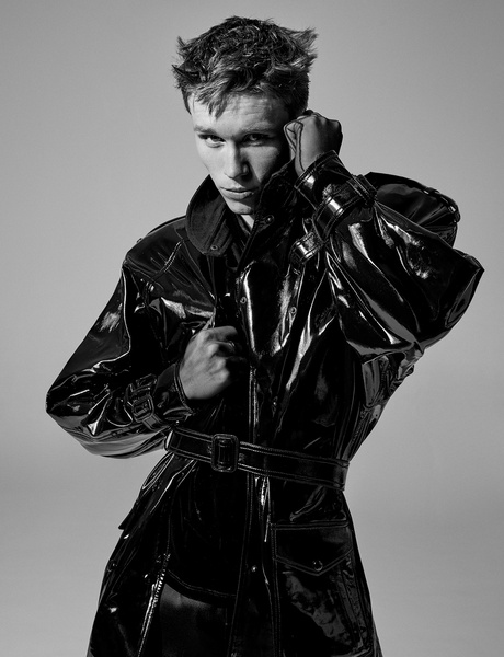 Monochrome portrait of a young adult male model in a shiny black trench coat, posing in a studio setting, with dramatic lighting.