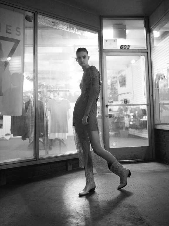 Monochrome image of a model wearing a patterned dress and cowboy boots standing in front of a storefront window in London.