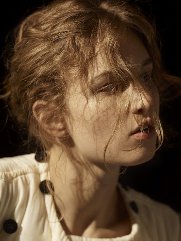 Portrait of a blonde woman in a light-colored top with dark buttons against a dark background, with soft lighting highlighting her face.