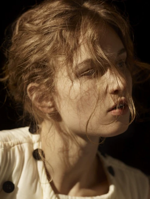 Portrait of a blonde woman in a light-colored top with dark buttons against a dark background, with soft lighting highlighting her face.