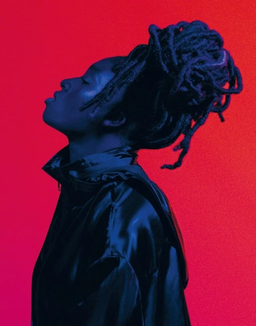 Side view of an adult with dreadlocks looking upwards, wearing a jacket in front of a red and pink gradient background in a studio.