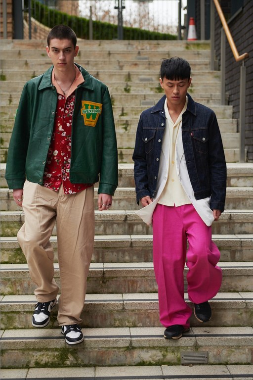 Two young men in fashionable, brightly colored wide-leg trousers walk down concrete steps in an urban environment, showcasing street style.