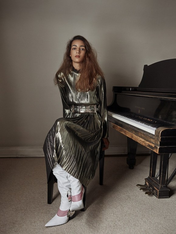 A woman with long brown hair in a shiny pleated gold dress sits on a stool in front of a black grand piano indoors.