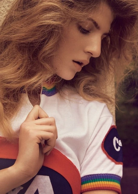 Close-up of a young woman with wavy auburn hair looking down, wearing a white t-shirt with rainbow trim and a red, white, and blue logo.
