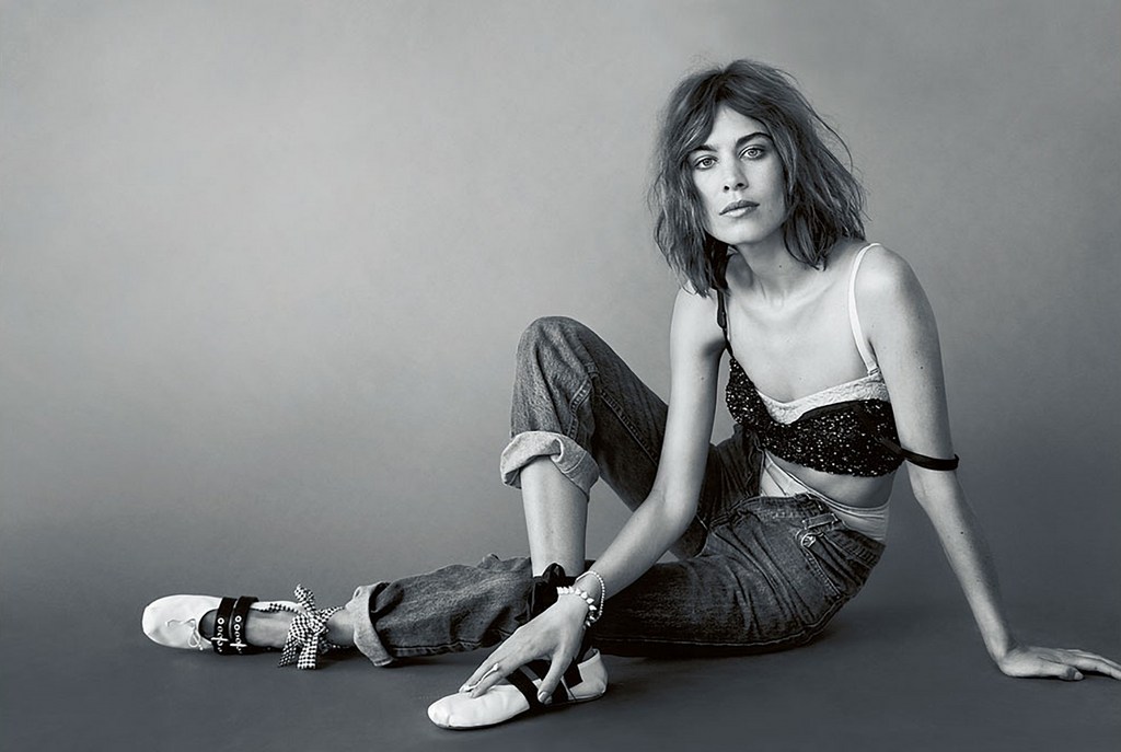 Black and white portrait of a young woman with wavy hair, wearing a bra top and jeans, posing in a photo studio.