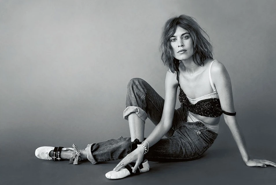 Black and white portrait of a young woman with wavy hair, wearing a bra top and jeans, posing in a photo studio.