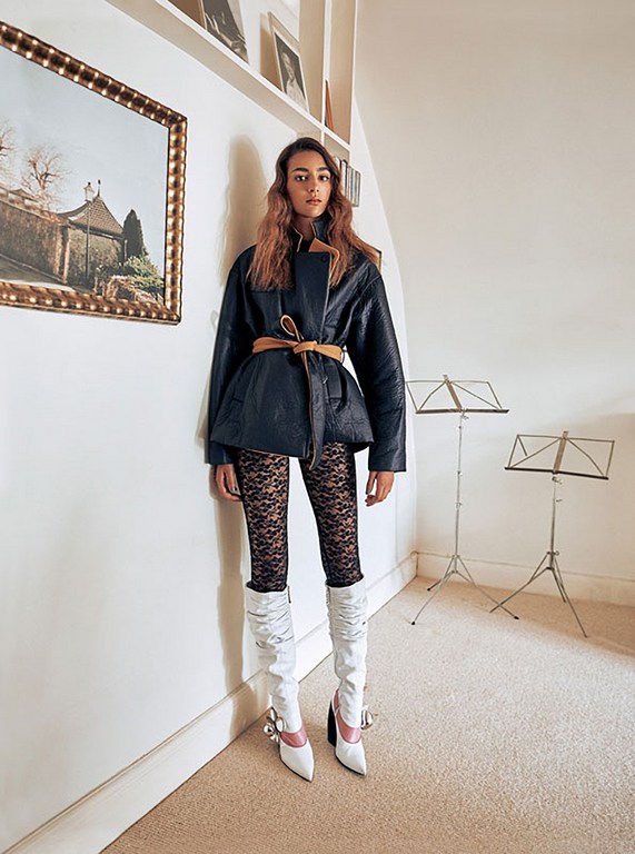 A woman in a black jacket, lace leggings, and white boots poses in an indoor setting with a painting and music stands.