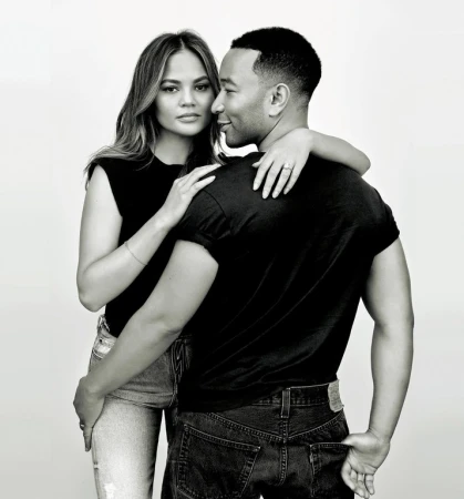 Black and white portrait of Chrissy Teigen and John Legend embracing in casual wear against a plain background in a studio setting.
